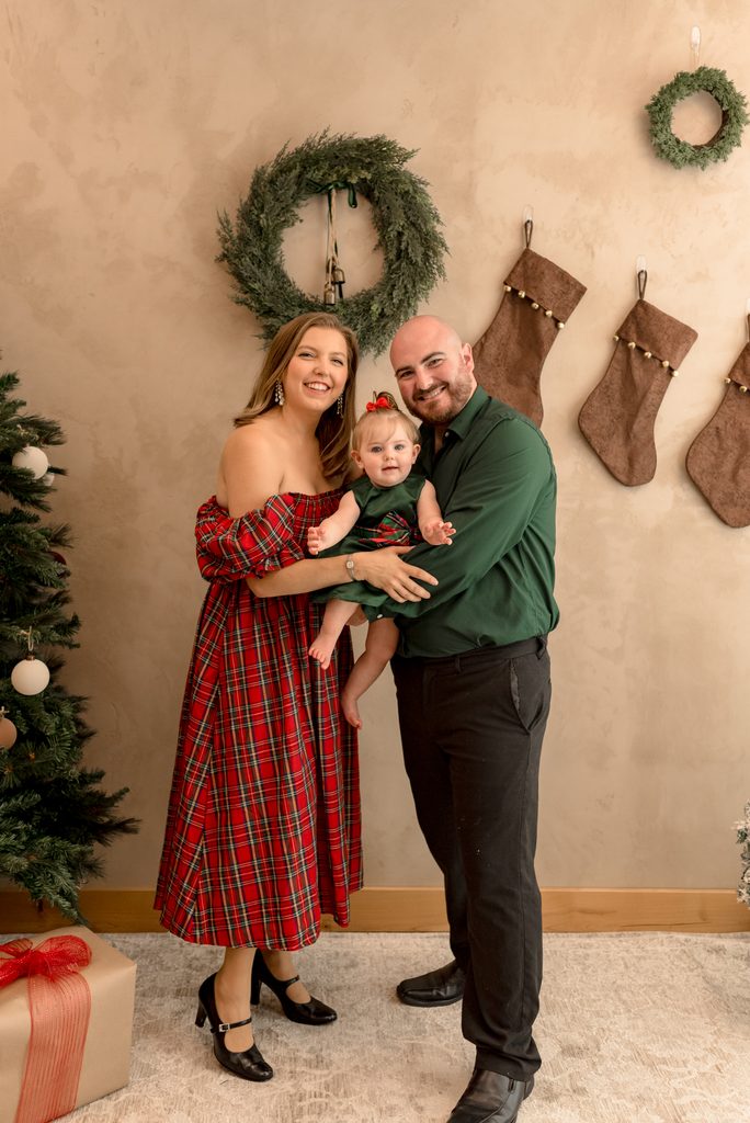 A mom, dad, and baby smile for the camera in front of a woodland themed Christmas studio background. 