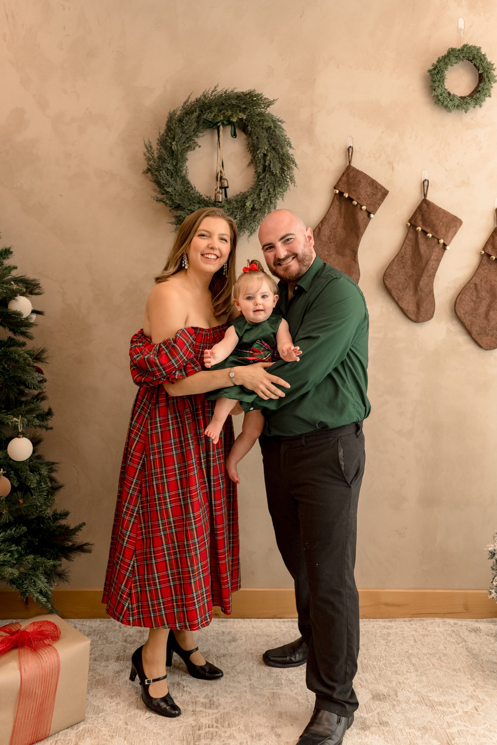 A young family stands in from of a Christmas wreath and stockings, smiling.