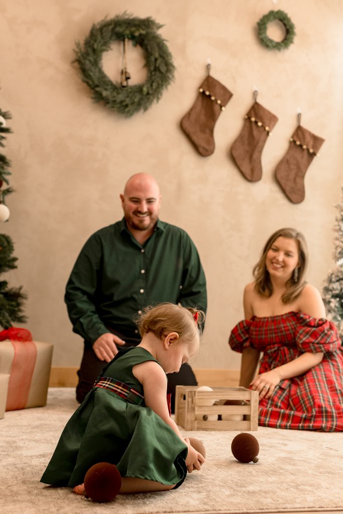A baby plays while her parents watch her with a Christmas wreath and stockings hanging in the background. 