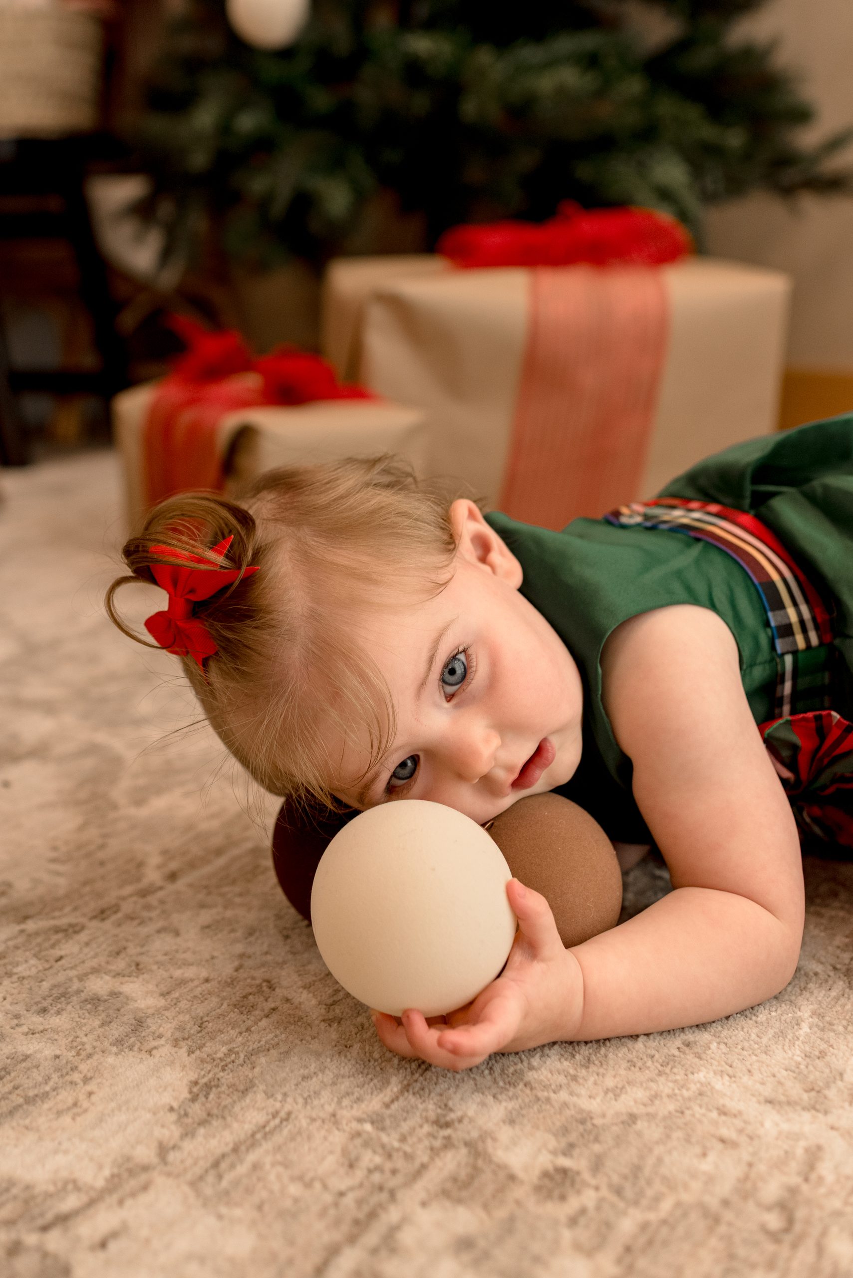 A baby holds Christmas bulbs with a tree in background at a studio mini session in Lebanon, Oregon. 