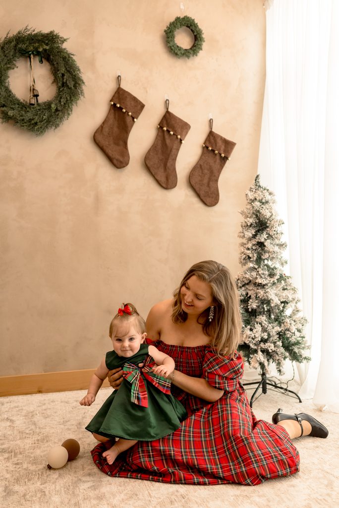 A mom sits in front of a Christmas tree with her daughter and smiles. 