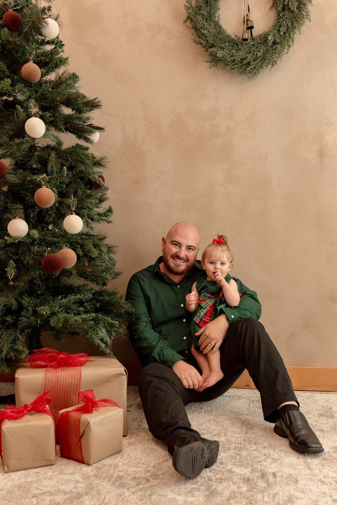 A dad sits beside a Christmas tree holding his baby at a studio photoshoot in Lebanon, Oregon. 